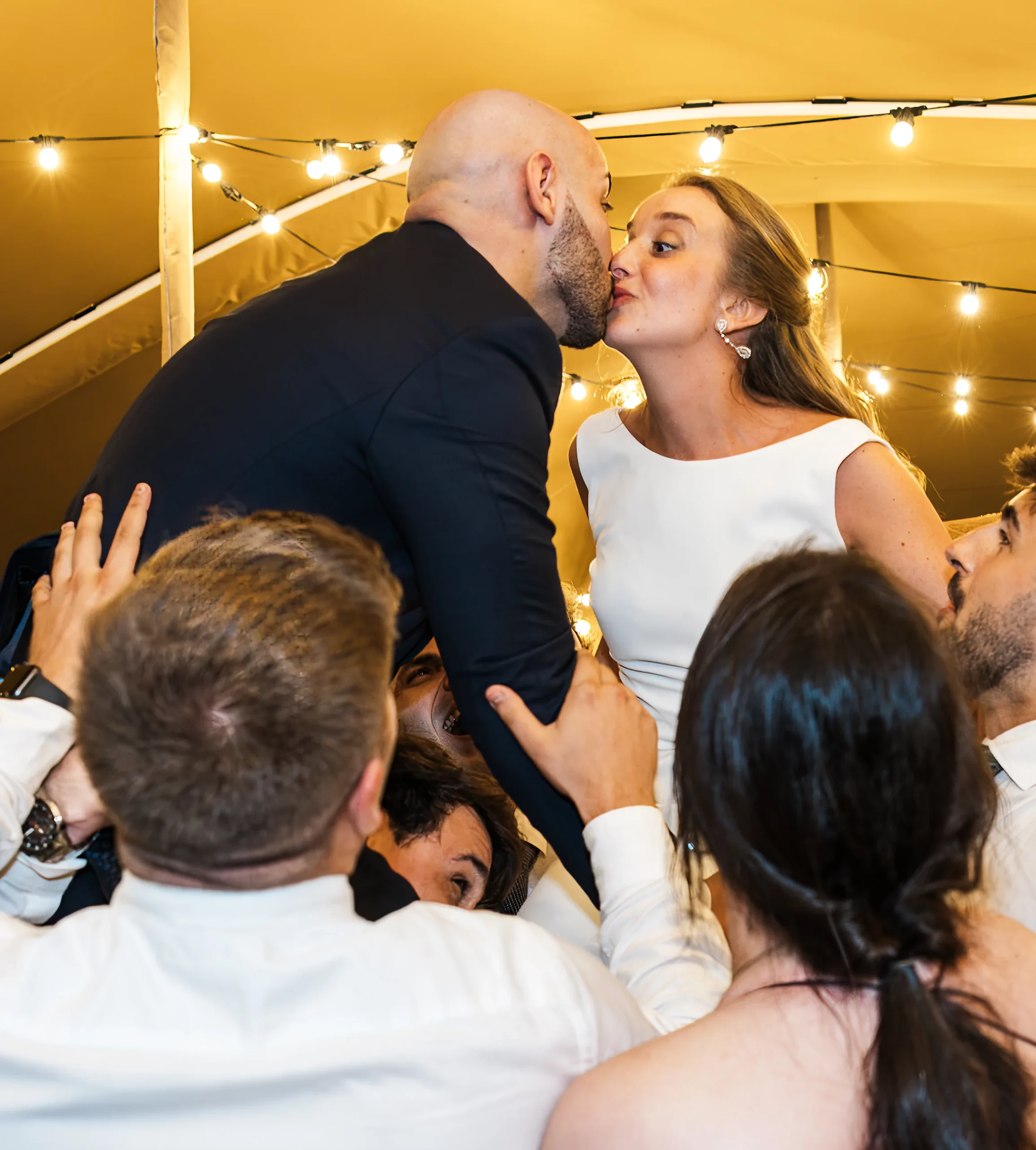 Novios dándose un beso durante la boda, levantados en el aire por los amigos, bajo una carpa iluminada con guirnaldas de luces.